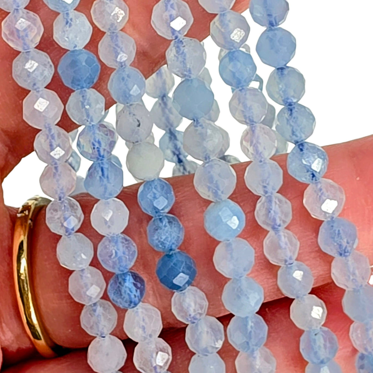 Faceted round gemstone beads in light blue and white, draped over a hand with a gold ring, on a white background.
