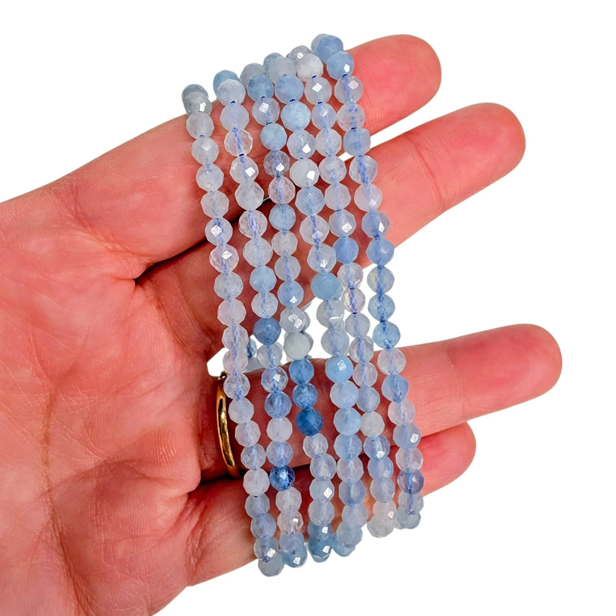 A hand holding five strands of faceted light blue beads, with a gold ring visible on one finger, against a plain white background.