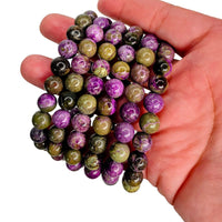A hand holding a bundle of round, polished gemstone beads in purple, olive green, and black with natural marbled patterns, set against a plain white background.