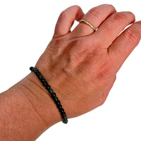 A close-up of a person's hand wearing a dark green beaded bracelet on the wrist and a thin gold ring on the finger, against a plain white background.