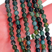Close-up of a hand holding several strands of round beads with engraved crescent moon symbols, showing a mix of green, blue-green, and dark navy stone colors.