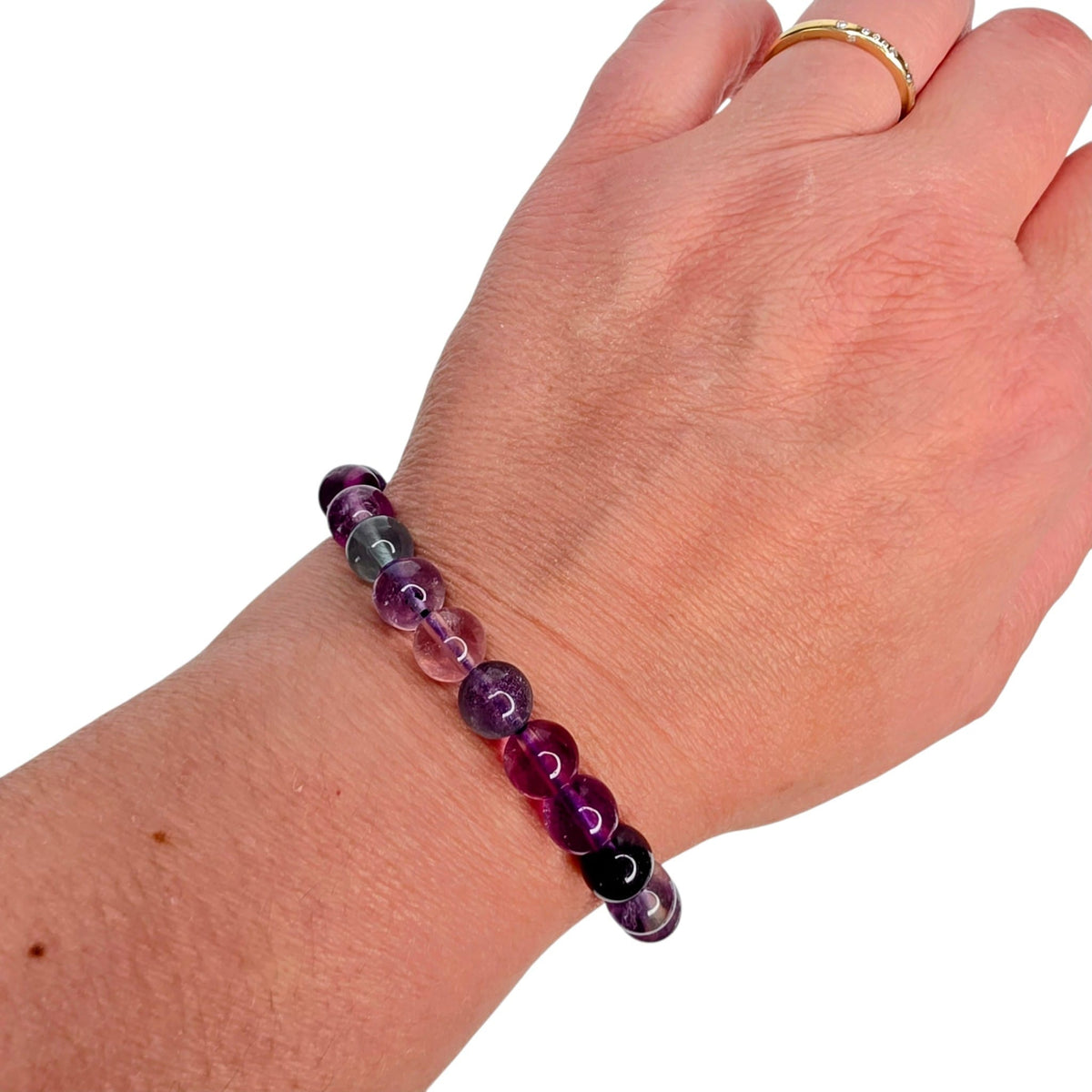 A beaded stretch bracelet made of polished purple, pink, and green gemstone beads worn on a person's wrist, with a gold ring on the finger, against a plain white background.
