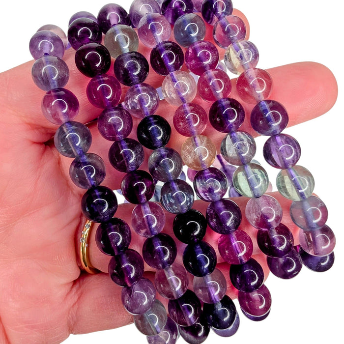 A hand holding several strands of polished round gemstone beads in shades of purple, violet, pink, and clear, with a gold ring visible on one finger against a white background.